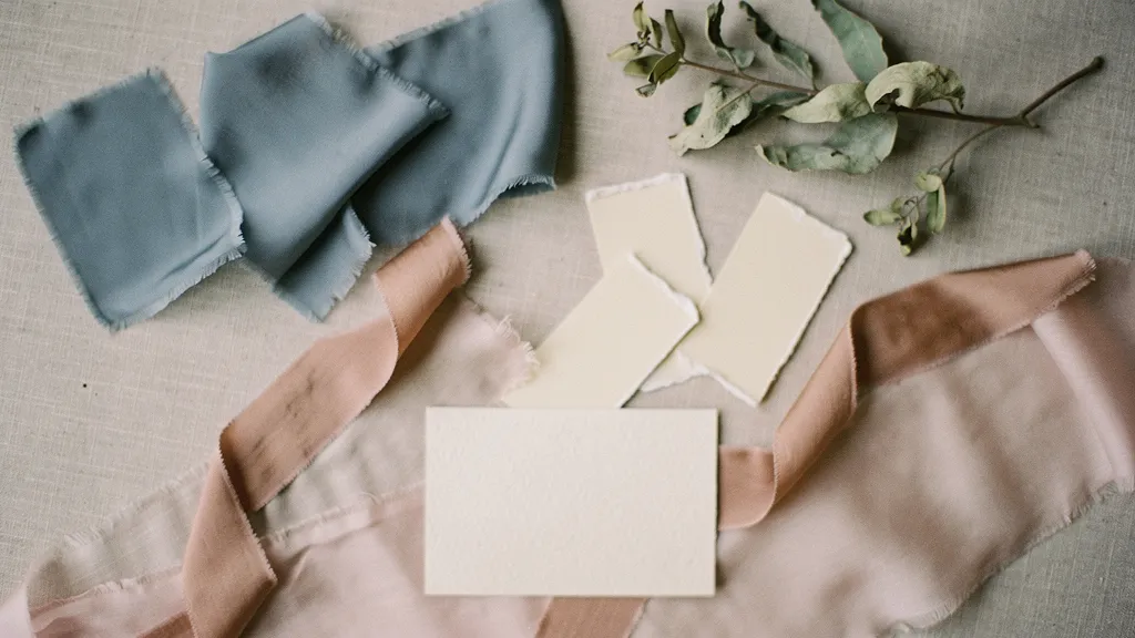 A carefully arranged wedding mood board with dusty blue fabrics, ivory paint chips, sage leaves, a taupe ribbon, and a blank card.