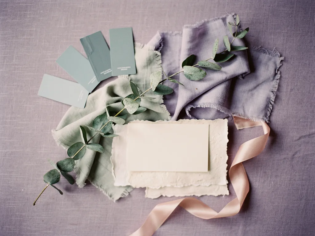 A wedding mood board features fabric swatches, eucalyptus leaves, paint chips, a ribbon, a blank card, and textured paper in soft, muted colors.