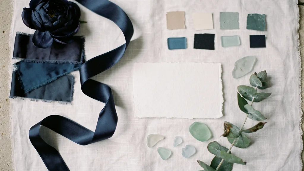 A wedding mood board with navy silk, white linen, sea glass, eucalyptus sprigs, paint chips, a navy ribbon, and a blank card.