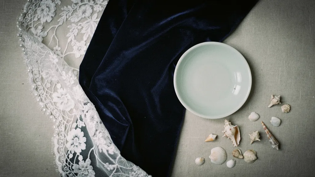 Navy velvet fabric, white lace, a sea foam ceramic dish, and small seashells arranged in an overhead view.